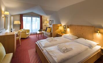 Camera matrimoniale Charme dell’Hotel St. Anton con letto in legno, angolo salotto e illuminazione accogliente.