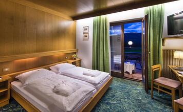 Camera matrimoniale Classic dell’Hotel St. Anton con letto in legno, scrivania e balcone con vista sulle montagne.