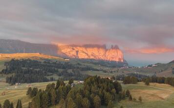 Schlern at sunset with green alpine meadows, forest, and low-hanging clouds.