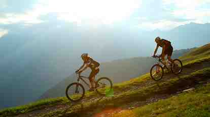 Two mountain bikers in a green alpine meadow at sunset; in the background, mountains and valleys.