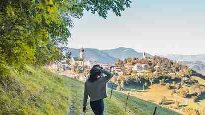 Woman on a meadow path with a view of a village and mountains.