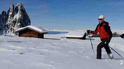 Due escursionisti con le ciaspole sull’Alpe di Siusi; baite in legno e un imponente massiccio montuoso sullo sfondo.