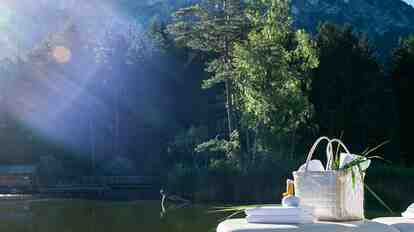 A woman in a white bathrobe sits by the Völser Weiher; in the background are forest, water and mountain scenery in the sunshine.