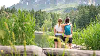 Two women stand among the reeds and look out over the Völser Weiher; forest and mountains in the background.