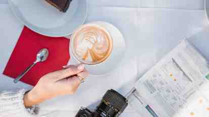 Tasse Cappuccino auf einem Tisch mit Kuchenstück, Kamera und aufgeschlagener Zeitung.