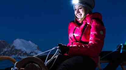 Nahaufnahme von zwei Nacht-Rodlern mit Stirnlampe; im Hintergrund verschneite Berge.
