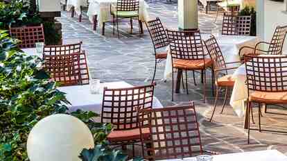 Sunny restaurant terrace with set tables, metal chairs, plants, and garden lights.