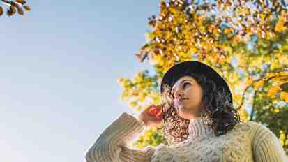 Woman with a hat and knitted sweater standing under a tree with autumn-colored leaves.