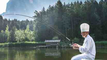 Cuoco in abbigliamento da cucina bianco pesca in un lago davanti a un bosco e a uno scenario alpino.