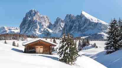 Baita in legno sull’Alpe di Siusi innevata; sullo sfondo il paesaggio montano invernale delle Dolomiti.