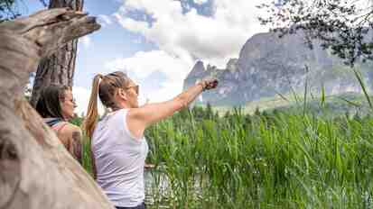 Two women sit among the reeds at the Völser Weiher, looking at the mountain backdrop of the Schlern.