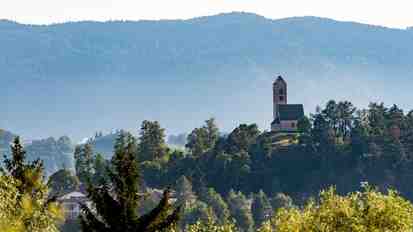 Church on a wooded hill in Völs am Schlern; in the background, a green mountain and forest landscape.