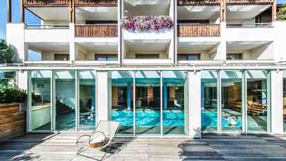 Vista esterna della piscina coperta dell’Hotel St. Anton con facciata in vetro, terrazza solarium e balconi decorati con fiori.