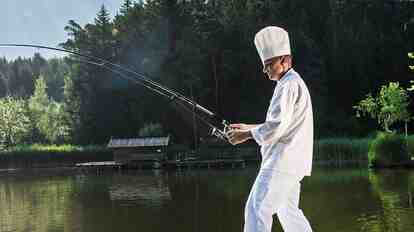 Cuoco in abbigliamento da cucina bianco pesca in un lago davanti a un bosco e a uno scenario alpino.