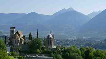 Merano with churches and green hillsides; in the background, the wide valley and the mountain scenery of South Tyrol.